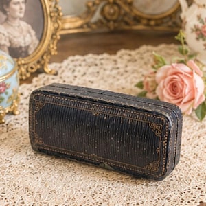 May include: A rectangular black jewellery box with a textured surface and gold-coloured decorative border detailing. The box features a hinged lid and a structured, elongated shape, showcasing a dark finish with subtle gold-toned line accents.