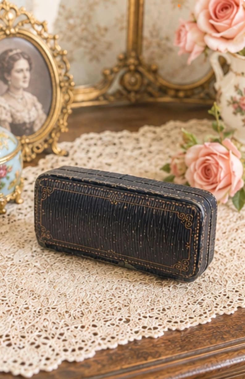 May include: A rectangular black jewellery box with a textured surface and gold-coloured decorative border detailing. The box features a hinged lid and a structured, elongated shape, showcasing a dark finish with subtle gold-toned line accents.