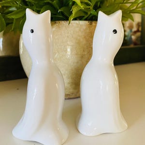 May include: Two white ceramic cat figurines with black eyes. The cat figurines have a sleek, minimalist design, with a curved body and pointed ears. The figurines are displayed on a white surface.