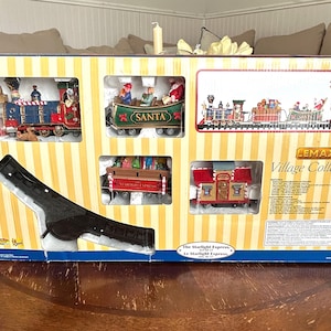 May include: A boxed Lemax Village Collection set featuring a Christmas train scene. The box has a yellow and white striped background with cutouts displaying miniature train cars, including a Santa-themed car and a red and blue train engine.