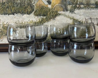 May include: A collection of eight gray-tinted, round drinking glasses. The glasses are stacked in pairs, showcasing their simple, elegant design. The glassware is made of a transparent material, allowing light to pass through.