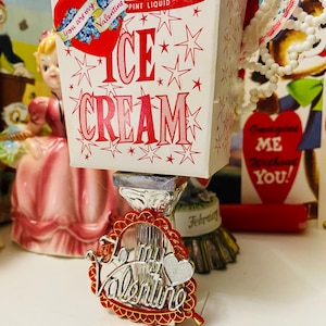 May include: A vintage-style ice cream container with red text and star accents, labeled "ICE CREAM" and "PINT LIQUID." A heart-shaped Valentine's Day pin with the words "Be my Valentine" is in front of the container. A small figurine is in the background.