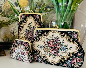 May include: Three vintage tapestry coin purses with floral designs. The purses vary in size, with gold-toned metal frames and clasps. The tapestry features a cream background with a black border and colourful flowers in shades of pink, red, and blue.