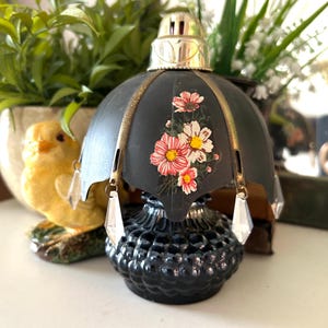 May include: Black glass perfume bottle with a decorative black and gold umbrella-shaped top. The top features a floral design with pink and white flowers. Clear crystal-like pendants hang from the umbrella.