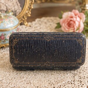 May include: A rectangular, dark navy blue jewellery box with a textured, rippled surface and gold-coloured embossed filigree detailing along the edges. The box features a hinged lid and a structured, elongated shape with rounded corners.