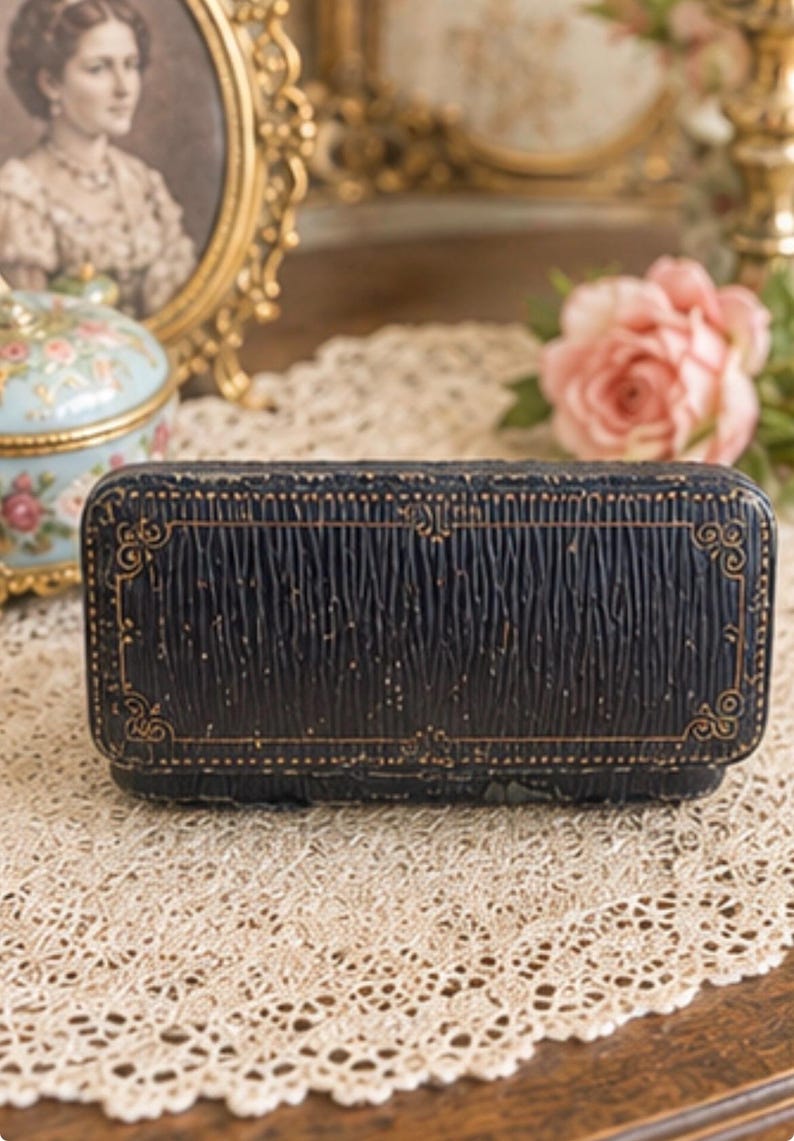May include: A rectangular, dark navy blue jewellery box with a textured, rippled surface and gold-coloured embossed filigree detailing along the edges. The box features a hinged lid and a structured, elongated shape with rounded corners.