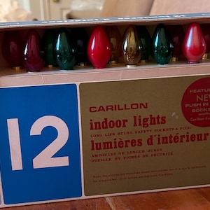 Peut inclure: Une boîte de 12 lumières d'intérieur Carillon avec des ampoules rouges, vertes et dorées. L'emballage est bleu et doré avec des motifs d'étoiles et le texte : 12 Carillon indoor lights, lumières d'intérieur, ampoules de longue durée.