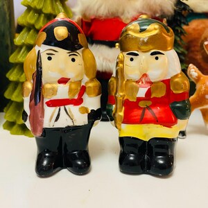 May include: Two vintage ceramic nutcracker figurines. One is dressed in black, white, and red with a black hat and gold accents. The other is in red, gold, and green with a gold crown. Both have painted faces and are part of a Christmas decoration collection.