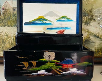 May include: Black lacquered jewellery box with a hinged lid and multiple compartments. The box features hand-painted Japanese-style landscape scenes with mountains, water, and a red building. The interior is lined with blue fabric.