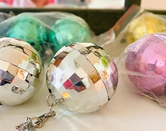 May include: Close-up of several disco ball ornaments in various colours, including silver, pink, green, and yellow. Each ball is covered in small, reflective mirrored tiles and attached to a silver chain. The ornaments are partially enclosed in clear plastic.