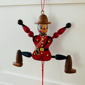 May include: A wooden jumping jack toy shaped like a soldier in a red uniform with a brown hat, black boots, and red arms. The figure features painted facial details, a yellow belt, and is suspended by a red string that controls the moving limbs.