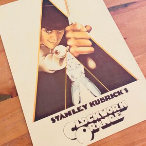 May include: A beige poster featuring the film "A Clockwork Orange." The poster has a central image of a person with a bowler hat, holding a knife and an eyeball. The film title and director's name are printed in bold, stylized lettering.