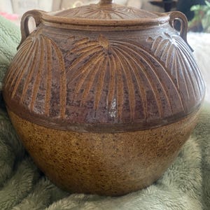 以下が含まれることがあります： 蓋付きの茶色の陶器の壺で、スタイリッシュな葉の彫刻が施されています。壺は両側に2つの取っ手があり、丸みを帯びた形をしています。