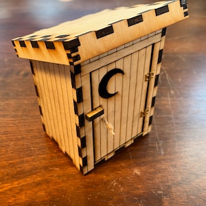 May include: A wooden outhouse with a black crescent moon on the door. The outhouse has a hinged door with a rope handle and a shingled roof.