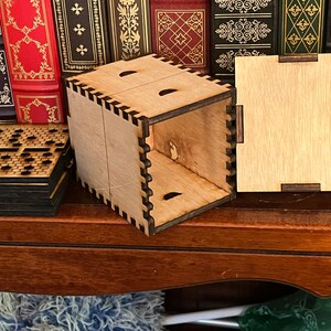 May include: A wooden box with a hinged lid, the box is open and the lid is lying flat on a wooden surface. The box is made of light wood with a dark brown stain. The lid has a simple design with a few small holes cut into it. The box is sitting on a wooden surface with a bookshelf in the background.