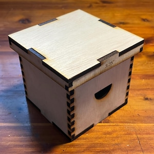 May include: A small, wooden box with a hinged lid. The box is made of light-colored wood with a dark brown trim. The lid has a small, rectangular cutout on the top. The box is labeled "3.25" on the side.