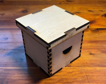 May include: A small, wooden box with a hinged lid. The box is made of light-colored wood with a dark brown trim. The lid has a small, rectangular cutout on the top. The box is labeled "3.25" on the side.
