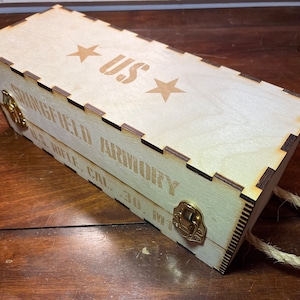 May include: A wooden box with a rope handle. The box is labeled "Springfield Armory, U.S. Rifle, Cal. .30, M1" and has a brass latch. The top of the box has two stars and the letters "US" in a stencil font.