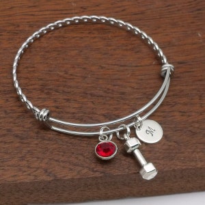 May include: Silver expandable bangle bracelet with a red crystal charm, a dumbbell charm, and a personalized charm with the letter "M".