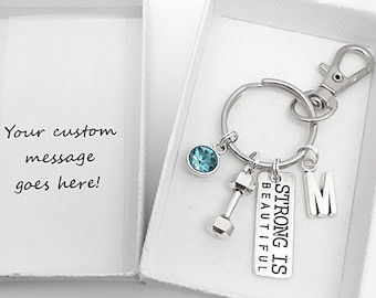 Op de afbeelding: Een zilveren sleutelhanger met een turquoise kristallen bedel, een halterbedel en een bedel met de tekst "Strong is Beautiful". De sleutelhanger heeft ook een zilveren letter "M" bedel en een zilveren sleutelring.