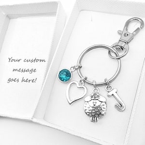 May include: A silver keyring with a charm of a sheep, a heart, a turquoise blue crystal, and a letter J. The keyring is in a white box with the text "Your custom message goes here!"