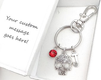 May include: A silver keychain with a sugar skull charm, a red crystal charm, and a letter J charm. The keychain is in a white gift box with the text "Your custom message goes here!"