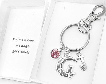 May include: A silver keychain with a crescent moon charm, a pink crystal charm, and a letter J charm. The keychain is in a white gift box.