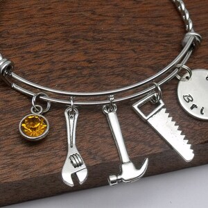 May include: A silver charm bracelet with a yellow crystal charm, a wrench charm, a hammer charm, a saw charm, and a round charm with the text "Bridget" and "a little" on it.