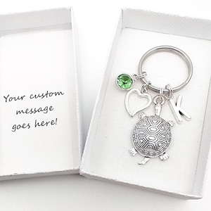 May include: A silver turtle keychain with a green crystal charm, a heart charm, and a letter K charm. The keychain is in a white gift box with a message that says "Your custom message goes here!"