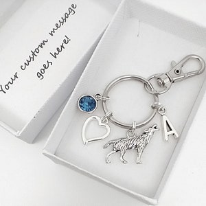 May include: A silver key ring with a wolf charm, a heart charm, a blue crystal charm, and a letter A charm. The key ring is in a white box with the text "Your custom message goes here!"