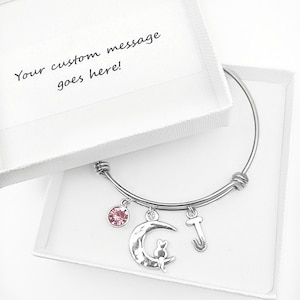 May include: A silver bangle bracelet with a charm of a crescent moon with a cat sitting on it, a pink crystal charm, and a letter J charm. The bracelet is in a white gift box with the text "Your custom message goes here!" on the inside.