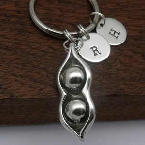 May include: Silver pea pod keyring with two peas inside. The keyring has two silver charms with the letters "R" and "H" on them.