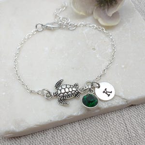 May include: A silver chain bracelet with a silver turtle charm, a green crystal charm, and a silver charm with the letter K.