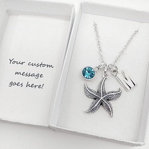May include: A silver necklace with a starfish charm, a blue crystal charm, and a silver charm with the letter "M". The necklace is in a white box with a message that says "Your custom message goes here!"