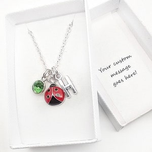 May include: A silver necklace with a ladybug charm, a green gemstone charm, and a silver letter H charm. The necklace is in a white gift box with a card that says "Your custom message goes here!"