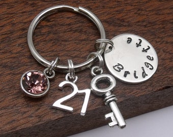May include: Silver keychain with a pink crystal, a key charm, a charm with the number 21, and a round charm with the word "Bridge" engraved on it.