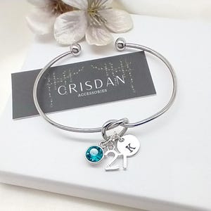 May include: Silver bangle bracelet with a knot design and charms. The charms include a turquoise crystal, a silver charm with the number 21, and a silver disc with the letter K.