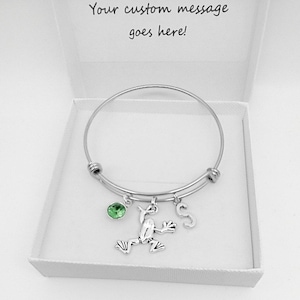 May include: A silver expandable bangle bracelet with a green crystal charm, a frog charm, and a letter 'S' charm. The bracelet is in a white box with the text 'Your custom message goes here!'