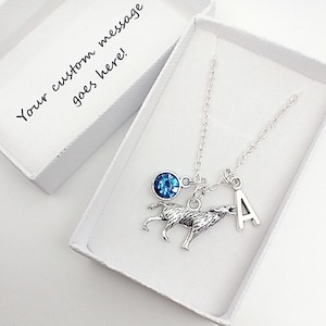 May include: A silver necklace with a blue crystal charm, a wolf charm, and a letter 'A' charm. The necklace is in a white gift box.