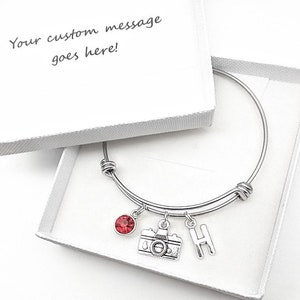 May include: A silver bangle bracelet with a red crystal charm, a camera charm, and a letter H charm. The bracelet is in a white box with the text "Your custom message goes here!"