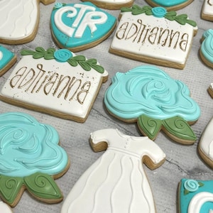 May include: Assortment of decorated sugar cookies. Some cookies are shaped like a dress, a rose, and a house with the name "Adrianna" in gold lettering. Other cookies are teal with white lettering.
