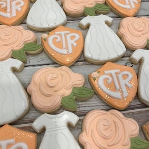 May include: Assortment of decorated sugar cookies. The cookies are shaped like white dresses, peach-colored roses with green leaves, and orange shields with the letters "CTR" in white. Some cookies have gold glitter accents.