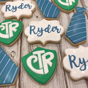 Könnte beinhalten: Auswahl an dekorierten Zuckerkeksen. Einige sind krawattenförmig mit blau-grünen Streifen. Andere sind weiß mit dem Namen "Ryder" in blauer Schrift. Einige sind grüne Schilde mit den Buchstaben "CTR" in Weiß.