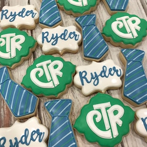 Puede incluir: Surtido de galletas de azúcar decoradas. Algunas tienen forma de corbata con rayas azules y verdes. Otras son blancas con el nombre "Ryder" en escritura azul. Las galletas verdes tienen "CTR" en blanco.
