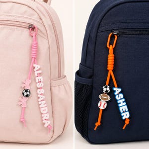May include: A light pink backpack with a pink cord keychain featuring the name ALESSANDRA, two pink bow charms, and a soccer ball bead, and a navy blue backpack with an orange cord keychain featuring the name ASHER, a soccer ball, football, and baseball bead.