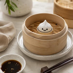 May include: A white dumpling with a smiling face sits in a bamboo steamer basket. The steamer is on a white plate, with a small bowl of sauce and chopsticks nearby. The scene is set on a neutral-toned surface.