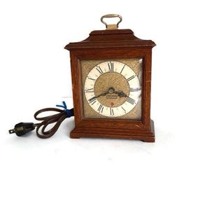 May include: A vintage wooden electric clock with a rectangular face and a handle on top. The clock face has Roman numerals and a gold-colored background. The clock is plugged into an electrical outlet.