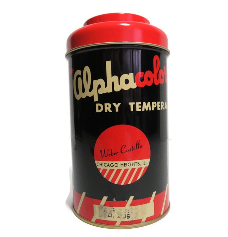 May include: A vintage tin container for Alphacolor Dry Tempera paint. The tin is black with red and gold lettering and a red lid. The label reads "Alphacolor Dry Tempera" and "Weber Costello, Chicago Heights, Ill."