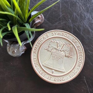 May include: A bronze-colored commemorative coin with the Liberty Bell at the center, surrounded by the text "America's Symbol of Freedom 1776-1976" and "Let It Ring." A small, decorative planter with green plants is in the upper left corner.