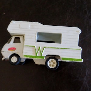 May include: A white toy camper van with green and red accents. The camper has a large window and a stylized "W" on the side. The wheels are black with white rims. A classic toy vehicle.